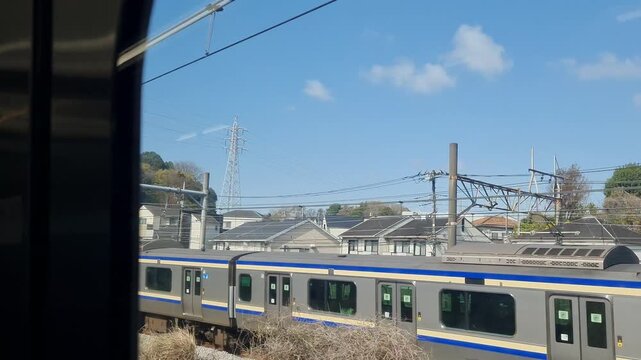 A modern Japanese commuter train travels through a suburban landscape with apartment buildings under a clear blue sky.