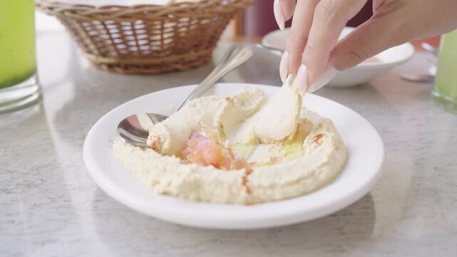 Close up of a female hand scooping creamy hummus with fresh pita bread from a white plate