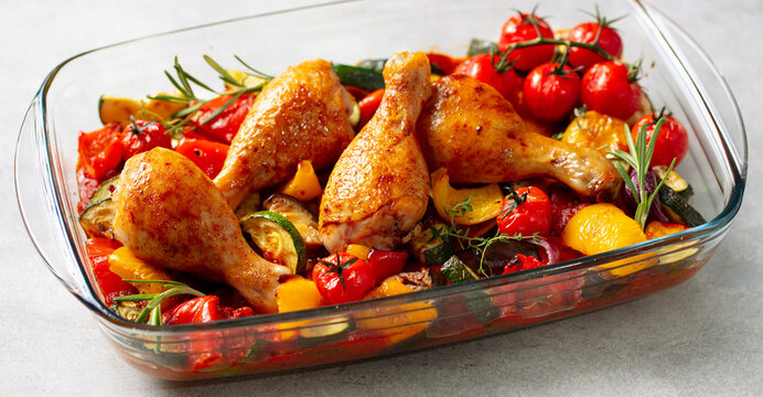 Baked chicken legs with zucchini and cherry tomatoes, garnished with parsley in glass baking dish. Grey background. Close up.