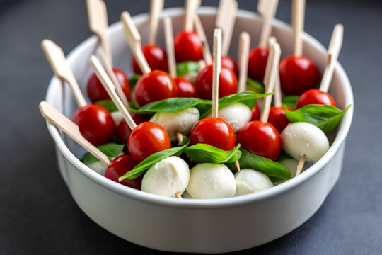 Fresh canape skewers with cherry tomatoes, mozzarella balls and basil leaves served in white bowl. The colorful appetizer reflects Italian caprese style, perfect for party snacks or festive catering