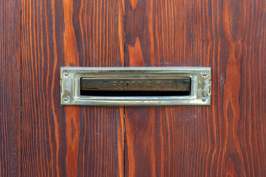 ​Horizontal close up photo of a vintage brass mailbox mail slot with Italian word Lettere installed on a natural reddish brown wooden door in daylight