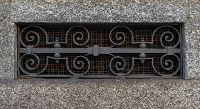 ​Horizontal photo of an ornate wrought iron basement window grille with scrollwork design set in a granite stone wall with protective wire mesh behind