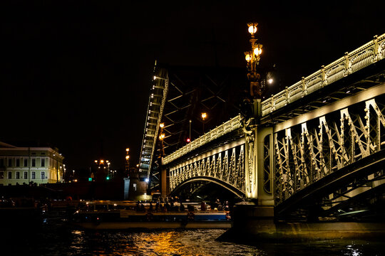 Trinity Bridge in St. Petersburg. Illuminated drawbridge at night with decorative street lamps and boats on the water, showcasing architectural details and urban scenery. Boat night cruise concept.