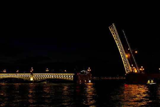 Trinity Bridge in St. Petersburg. Illuminated drawbridge with a raised section spans over dark water, reflecting city lights at night, highlighting the architectural design and urban landscape