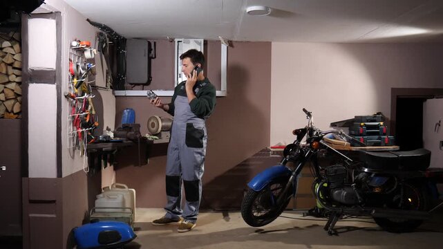 Man in overalls holding a mechanical part and talking on his smartphone in a home workshop, planning a repair or maintenance for a vintage motorcycle. He is surrounded by tools and equipment