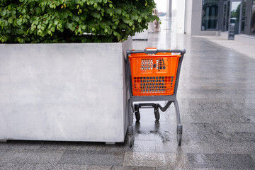 Empty orange shopping cart standing beside a large concrete planter on a wet pavement outside a modern commercial building. © kivitimof