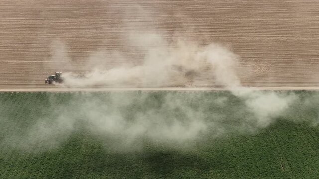 Farm tractor plowing dry agricultural field, creating huge dust cloud next to a green crop plot