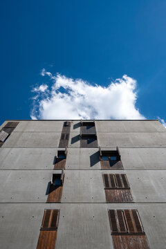 Against a blue sky, an upward view reveals concrete residential walls in La Confluence, where timber shutters and windows align beneath a cloud in Lyon, France