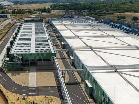 Aerial view of a modern data center facility with large white rooftops, green industrial facades, and surrounding access roads in the United Kingdom.
