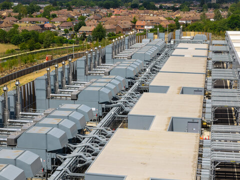 Aerial view of a data center rooftop featuring rows of industrial cooling units and ventilation systems with a suburban residential area in the background in the United Kingdom.