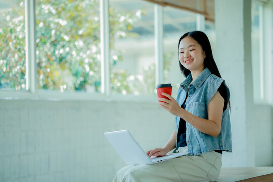 Young Asian woman reading a book with headphones and laptop by the window