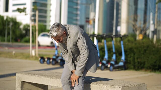 Mature Brazilian businessman in gray plaid suit touching knee in discomfort after standing from urban bench in city