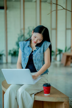Young Asian woman reading a book with headphones and laptop by the window