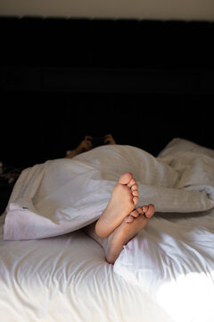Close-up of bare woman's feet sticking out from a cozy white duvet on a bed. Concept of a lazy morning, deep sleep, relaxation, comfort, and weekend leisure at home or a hotel room.