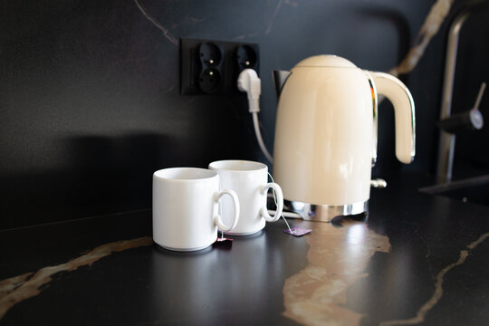 Elegant white electric kettle standing next to two ceramic mugs with tea bags on a dark kitchen tabletop. Cozy morning tea break, domestic appliance, and modern kitchen interior concept.