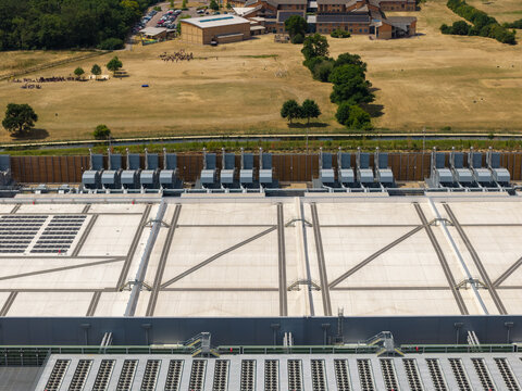 Aerial view of a large data center facility featuring extensive rooftop cooling units and HVAC systems, set against a backdrop of open fields and trees in the United Kingdom.