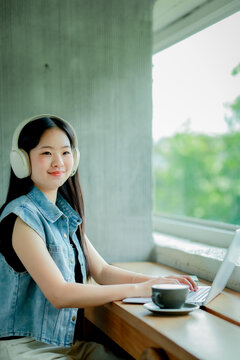 Young Asian woman reading a book with headphones and laptop by the window