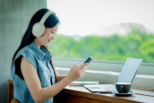 Young Asian woman talking on smartphone while working on laptop in modern workspace