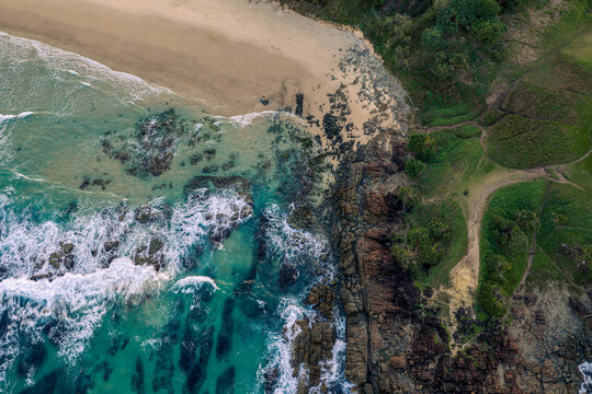 Aerial view of the rugged coastline where turquoise ocean waves meet sandy beaches and grassy cliffs in Byron Bay, New South Wales, Australia.