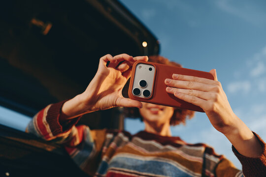 Woman lifestyle outdoors in daylight, taking a smartphone selfie with hand up, striped sweater, car background softly blurred, relaxed joyful mood, technology sharing, social media, smile