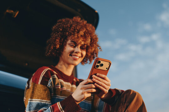 Woman lifestyle portrait outdoors, curly hair woman smiling while holding smartphone beside a car, casual striped sweater, daylight and blue sky background, relaxed mood for modern communication