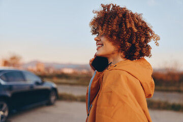 Woman lifestyle profile portrait, curly hair, smiling outdoors, wearing orange jacket, sunset light, soft background with shallow depth of field, parked cars nearby, casual street travel moment © SHOTPRIME STUDIO