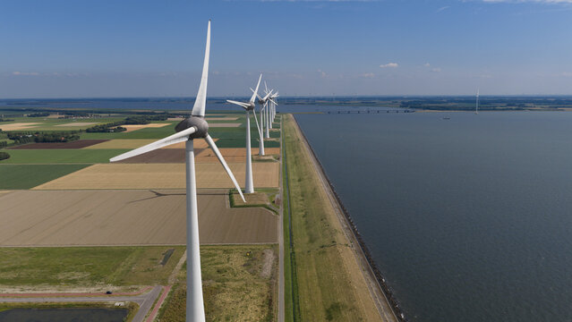 Aerial view of a row of wind turbines standing along a dike that separates agricultural fields from the water in Nagele, Flevoland, Netherlands.