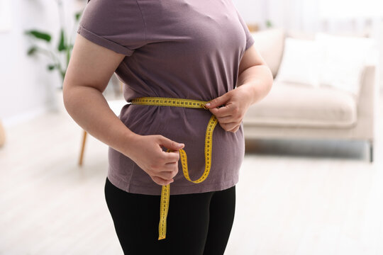 Weight loss. Overweight woman measuring her waist with tape indoors, closeup
