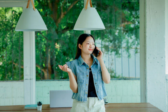 Young Asian woman talking on smartphone while working on laptop in modern workspace