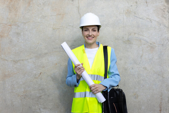 Woman architect engineer wearing a hard hat and safety vest, holding blueprints, looking at camera and smiling