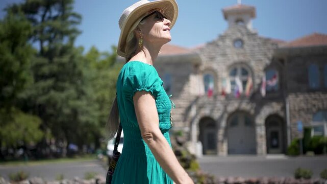 A senior woman smiles and enjoys a walk by City Hall in Sonoma California during a sunny day while surrounded by trees and flowers.