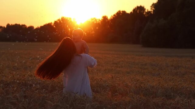 A happy mother and her seven-month-old son spin in a wheat field in the rays of the setting sun against a backdrop of soap bubbles