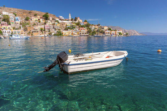 Small motor boat anchored in the bay with the colorful waterfront of Symi Island, Dodecanese, Greece, in the background.