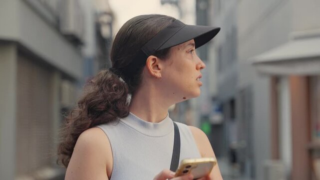Disoriented young woman wearing sun visor using her smartphone for navigation, looking around with a puzzled expression while trying to find her way through a narrow alley in a foreign city