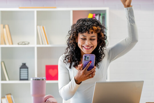 Smiling young woman with curly hair celebrating good news while looking at her smartphone, sitting at a desk with an open laptop in a bright home office