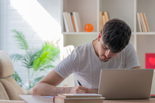 Young man writing notes in a notebook while working on a laptop at a home desk, focused on studying or freelance work