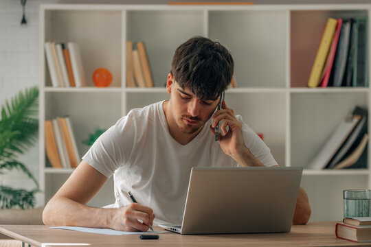 Young man multitasking at home office desk, talking on smartphone while taking notes on paper with laptop open in front of him, focused on work call
