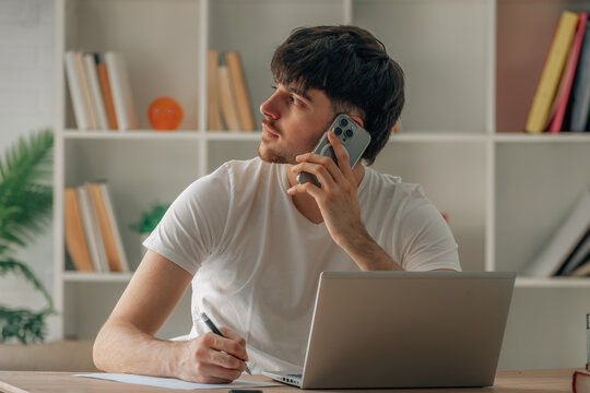 Young man multitasking at home office desk, talking on smartphone while taking notes on paper with laptop open in front of him, focused on work call