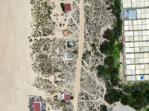 Aerial view of Praia da Mata showing sandy dunes, a dirt path, small buildings, and rows of white-roofed structures in Costa de Caparica, Setubal, Portugal.