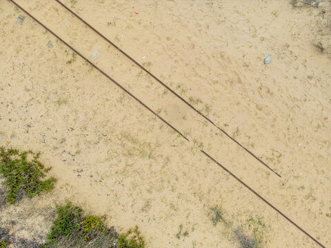 Aerial view of Praia da Mata featuring parallel rusted metal tracks running across the sandy terrain with sparse coastal vegetation in Costa de Caparica, Setubal, Portugal.