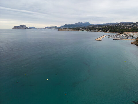Aerial view of the marina and coastline of Moraira, with calm turquoise waters and the Penon de Ifach visible in the distance under a cloudy sky in Moraira, Valencian Community, Spain.