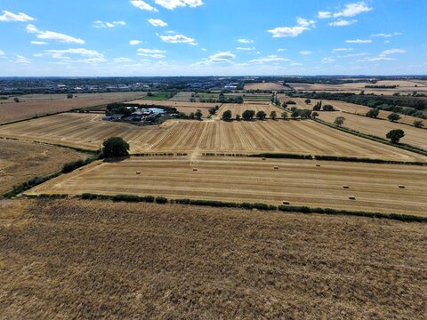 Aerial view of harvested wheat fields with scattered hay bales, green hedgerows, and farm buildings under a bright blue sky in Finedon, England, United Kingdom.