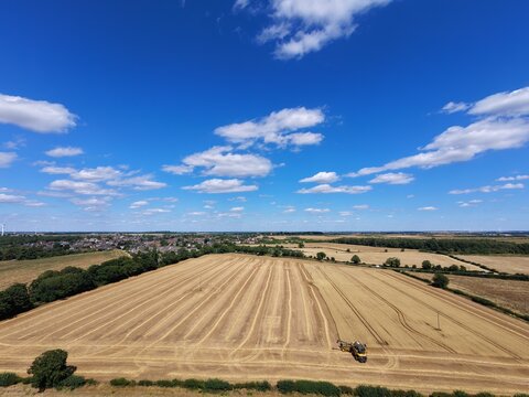 Aerial view of a combine harvester in a harvested wheat field with rows of straw and rural landscape in Finedon, England, United Kingdom.