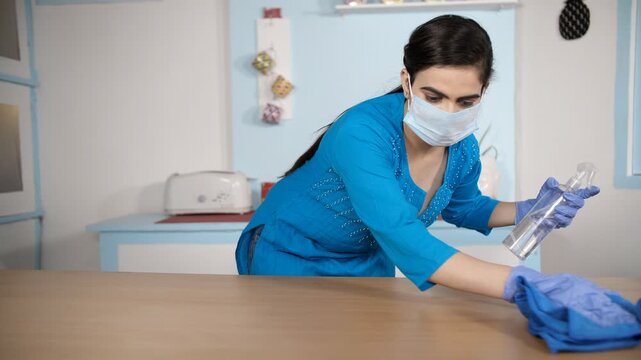 Young housewife taking care of hygiene / sanitizing to disinfect her home. Woman wearing gloves and mask cleaning workplace / table with an antibacterial wipe - Coronavirus protection concept