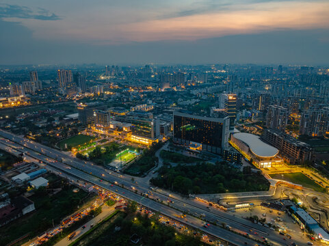 Aerial view of the modern cityscape at dusk, featuring illuminated skyscrapers, office buildings, and a busy highway with light trails from traffic in Gurgaon, Haryana, India.