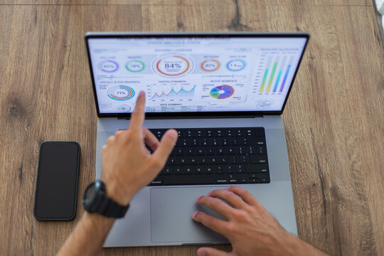 Overhead view of a businessman's hands analyzing financial graphs on a laptop screen, pointing a finger at data metrics. Smartphone on a wooden desk workspace.