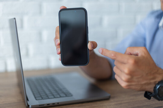 Close-up of a man holding a black smartphone with a blank mockup screen towards the camera and pointing at it. Laptop on a wooden desk, white brick wall background. Shallow depth of field.
