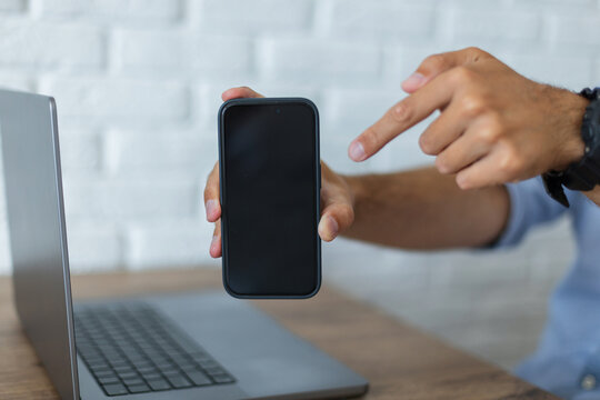 Close-up of a person holding a smartphone with a blank mockup screen, pointing a finger at it. Laptop on a wooden desk with a white brick wall background. Shallow depth of field.