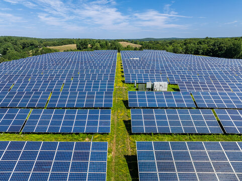 Aerial view of a large solar photovoltaic power plant with rows of solar panels on a capped landfill lined by trees, under a blue sky. Massachusetts, USA.