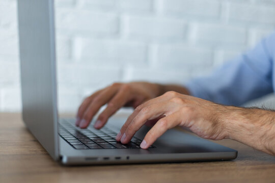 Close-up side view of a man's hands typing on a laptop keyboard. Working at a wooden desk in a bright office with a white brick wall background. Selective focus on hands.